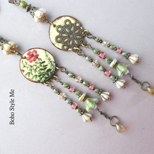 May include: A pair of long dangle earrings featuring circular components. One earring displays a floral print with a red flower, while the other features an intricate bronze filigree pattern. Both include beaded drapes with green, pink, and white accents.
