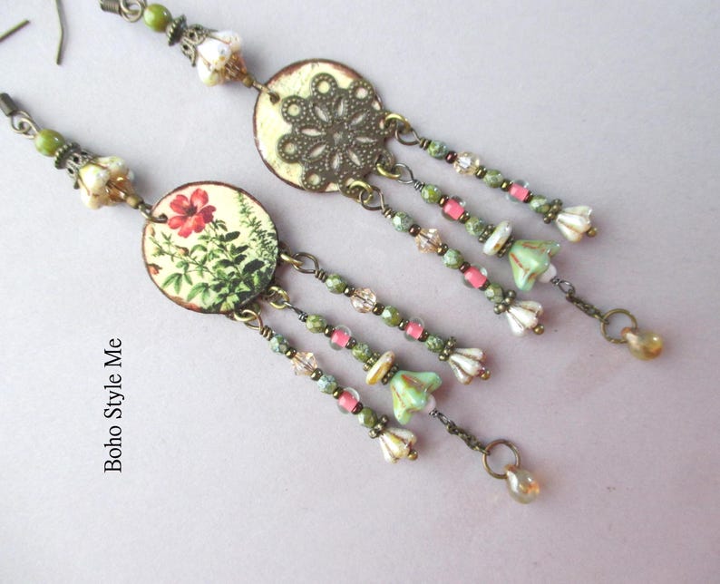 May include: A pair of long dangle earrings featuring circular components. One earring displays a floral print with a red flower, while the other features an intricate bronze filigree pattern. Both include beaded drapes with green, pink, and white accents.