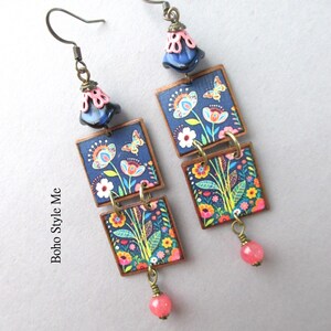 Puede incluir: Un par de pendientes colgantes con dos colgantes cuadrados sobre fondo azul oscuro con un estampado colorido de flores y mariposas. Cada pendiente tiene una cuenta azul oscuro en forma de campana con una tapa rosa y una cuenta rosa.