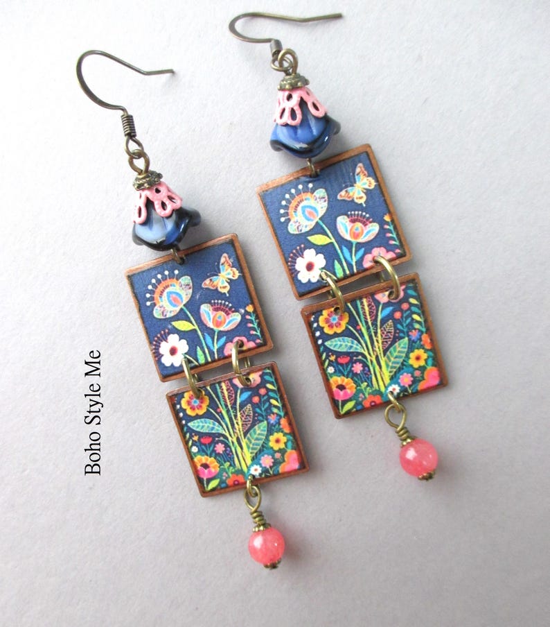 Puede incluir: Un par de pendientes colgantes con dos colgantes cuadrados sobre fondo azul oscuro con un estampado colorido de flores y mariposas. Cada pendiente tiene una cuenta azul oscuro en forma de campana con una tapa rosa y una cuenta rosa.