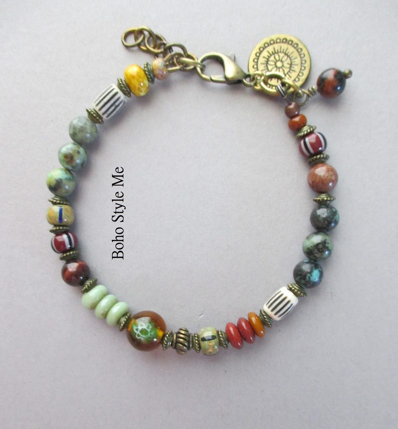 K&ouml;nnte beinhalten: Ein Armband mit einer Vielzahl von bunten Perlen, darunter gr&uuml;ne, braune, rote und wei&szlig; gestreifte Muster, akzentuiert mit antikfarbenen Messing-Abstandshaltern und einem runden Anh&auml;nger. Der Text "Boho Style Me" steht nahe der Mitte des Armbands.
