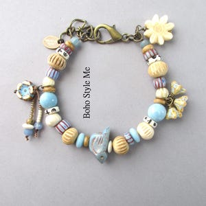 May include: A bracelet featuring a mix of light blue, cream, and striped beads, a central bird-shaped charm, and floral accents. The piece includes a bronze-toned lobster clasp and a small oval tag with the text Boho Style Me.
