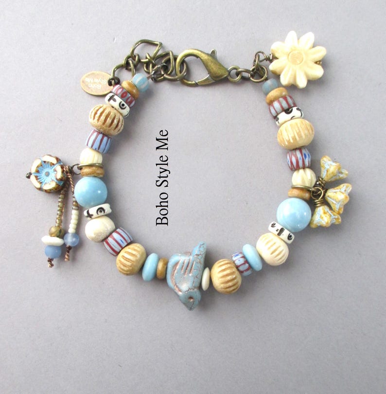 May include: A bracelet featuring a mix of light blue, cream, and striped beads, a central bird-shaped charm, and floral accents. The piece includes a bronze-toned lobster clasp and a small oval tag with the text Boho Style Me.