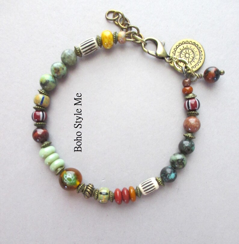 K&ouml;nnte beinhalten: Ein Perlenarmband mit einer Vielzahl von bunten Stein-, Glas- und gemusterten Perlen, die durch kleine bronzefarbene Metallabstandshalter getrennt sind. Das Armband verf&uuml;gt &uuml;ber einen Karabinerverschluss, eine Verl&auml;ngerungskette und einen runden Anh&auml;nger mit Sonnenmotiv.