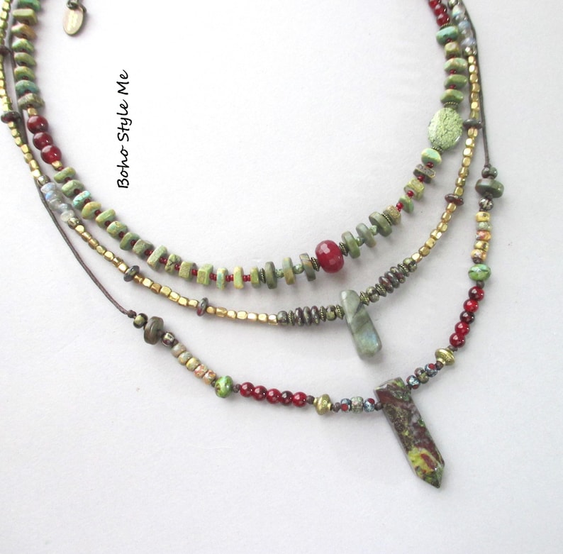 Puede incluir: Un collar de varias hileras con cuentas verdes y rojas, detalles en oro met&aacute;lico y dos colgantes de piedra. El texto "Boho Style Me" est&aacute; impreso en el lado izquierdo. El dise&ntilde;o incorpora varias cuentas peque&ntilde;as y texturizadas en cord&oacute;n oscuro.