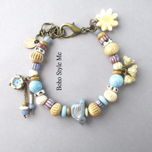 May include: A charm bracelet featuring a variety of beads in shades of blue, cream, and tan, including a central bird-shaped bead, a flower charm, and a lobster clasp. The text "Boho Style Me" is printed in the center of the bracelet loop.