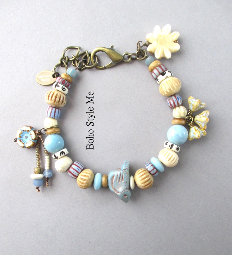 May include: A charm bracelet featuring a variety of beads in shades of blue, cream, and tan, including a central bird-shaped bead, a flower charm, and a lobster clasp. The text "Boho Style Me" is printed in the center of the bracelet loop.