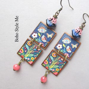 Puede incluir: Un par de pendientes colgantes con dos piezas cuadradas con estampado floral y de mariposas sobre fondo azul oscuro, acentuados por una cuenta rosa en la parte inferior y una cuenta azul con tapa rosa en la parte superior. El texto Boho Style Me es visible.
