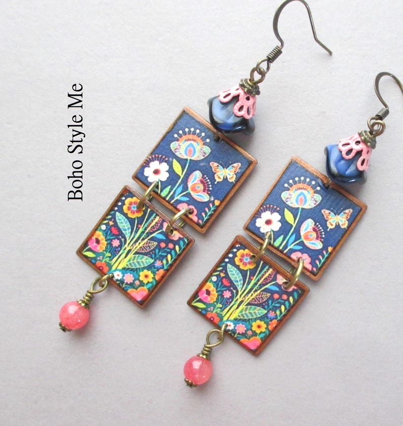 Puede incluir: Un par de pendientes colgantes con dos piezas cuadradas con estampado floral y de mariposas sobre fondo azul oscuro, acentuados por una cuenta rosa en la parte inferior y una cuenta azul con tapa rosa en la parte superior. El texto Boho Style Me es visible.