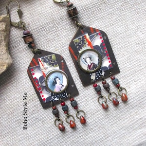 Puede incluir: Un par de pendientes colgantes con un marco de metal en forma de casa y un estampado de collage de una persona con sombrilla. Los pendientes incluyen herrajes de tono bronce, pequeñas cuentas oscuras y tres dijes colgantes con detalles rojos y grises.