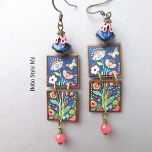 Puede incluir: Un par de pendientes colgantes con dos piezas cuadradas apiladas sobre un fondo azul oscuro con un estampado colorido de flores y mariposas. Cada pendiente tiene una cuenta azul y rosa en forma de campana y una peque&ntilde;a cuenta rosa en la base.