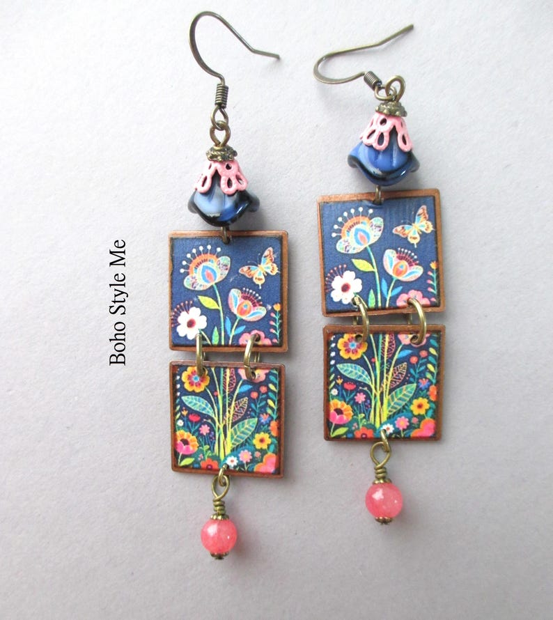 Puede incluir: Un par de pendientes colgantes con dos piezas cuadradas apiladas sobre un fondo azul oscuro con un estampado colorido de flores y mariposas. Cada pendiente tiene una cuenta azul y rosa en forma de campana y una peque&ntilde;a cuenta rosa en la base.