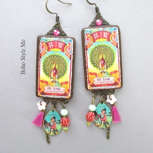 May include: A pair of rectangular drop earrings featuring a vintage Po Sing matchbox label with a peacock illustration. The earrings include bronze-toned metal findings, a pink rhinestone, small white flower beads, red striped beads, and pink tassels.