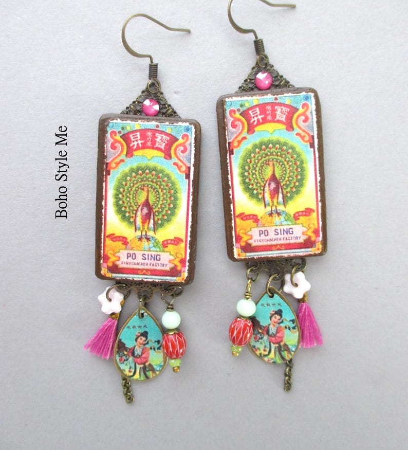 May include: A pair of rectangular drop earrings featuring a vintage Po Sing matchbox label with a peacock illustration. The earrings include bronze-toned metal findings, a pink rhinestone, small white flower beads, red striped beads, and pink tassels.