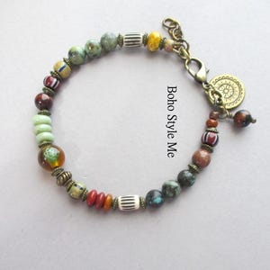 K&ouml;nnte beinhalten: Ein Perlenarmband mit runden, zylindrischen und scheibenf&ouml;rmigen Perlen in Gr&uuml;n, Rot, Braun, Gelb und Wei&szlig;, akzentuiert durch bronzefarbene Zwischenelemente, einen Karabinerverschluss und einen runden Anh&auml;nger mit Sonnenmotiv.