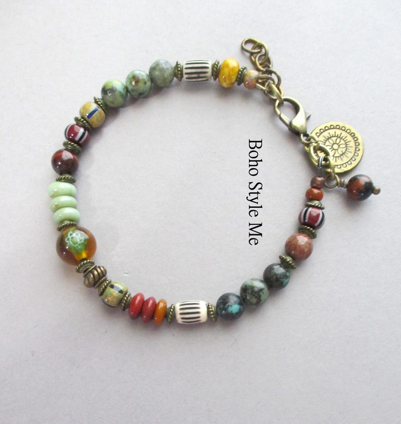 K&ouml;nnte beinhalten: Ein Perlenarmband mit runden, zylindrischen und scheibenf&ouml;rmigen Perlen in Gr&uuml;n, Rot, Braun, Gelb und Wei&szlig;, akzentuiert durch bronzefarbene Zwischenelemente, einen Karabinerverschluss und einen runden Anh&auml;nger mit Sonnenmotiv.