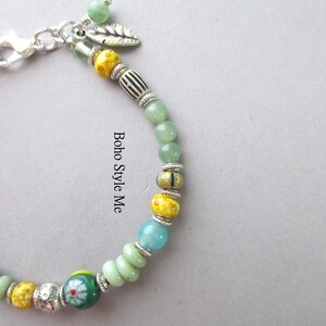 May include: A beaded bracelet featuring a variety of round and cylindrical beads in shades of green, yellow, and blue, accented with silver-toned spacers and a leaf charm. The text "Boho Style Me" is printed vertically along the center of the bracelet.