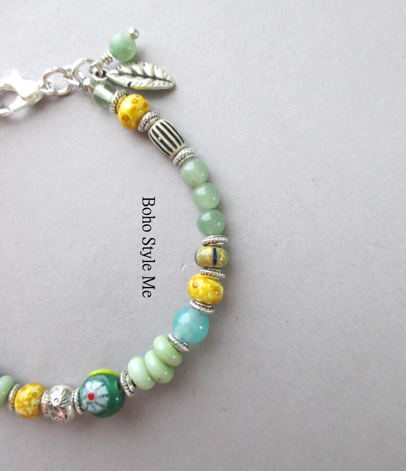 May include: A beaded bracelet featuring a variety of round and cylindrical beads in shades of green, yellow, and blue, accented with silver-toned spacers and a leaf charm. The text "Boho Style Me" is printed vertically along the center of the bracelet.