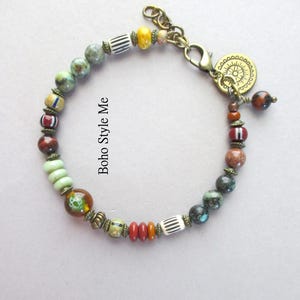 K&ouml;nnte beinhalten: Ein Perlenarmband mit einer Vielzahl von mehrfarbigen Stein-, Glas- und gestreiften Perlen, die durch kleine bronzefarbene Metallabstandshalter getrennt sind. Das Armband verf&uuml;gt &uuml;ber einen bronzefarbenen Karabinerverschluss und einen runden Anh&auml;nger.