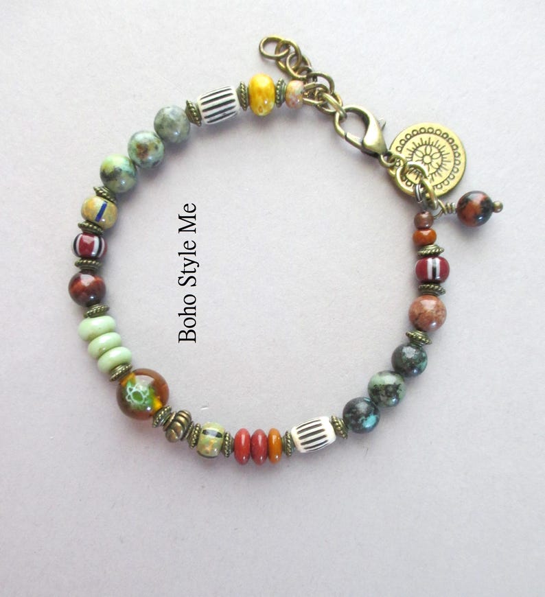 K&ouml;nnte beinhalten: Ein Perlenarmband mit einer Vielzahl von mehrfarbigen Stein-, Glas- und gestreiften Perlen, die durch kleine bronzefarbene Metallabstandshalter getrennt sind. Das Armband verf&uuml;gt &uuml;ber einen bronzefarbenen Karabinerverschluss und einen runden Anh&auml;nger.