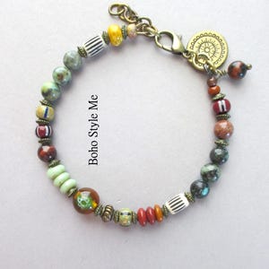 K&ouml;nnte beinhalten: Ein Perlenarmband mit einer Vielzahl bunter Perlen in Gr&uuml;n-, Braun-, Rot- und Gelbt&ouml;nen, akzentuiert durch antike Messing-Abstandshalter und einen runden Anh&auml;nger. Der Text Boho Style Me ist sichtbar. Es enth&auml;lt einen Karabinerverschluss und eine Kette.