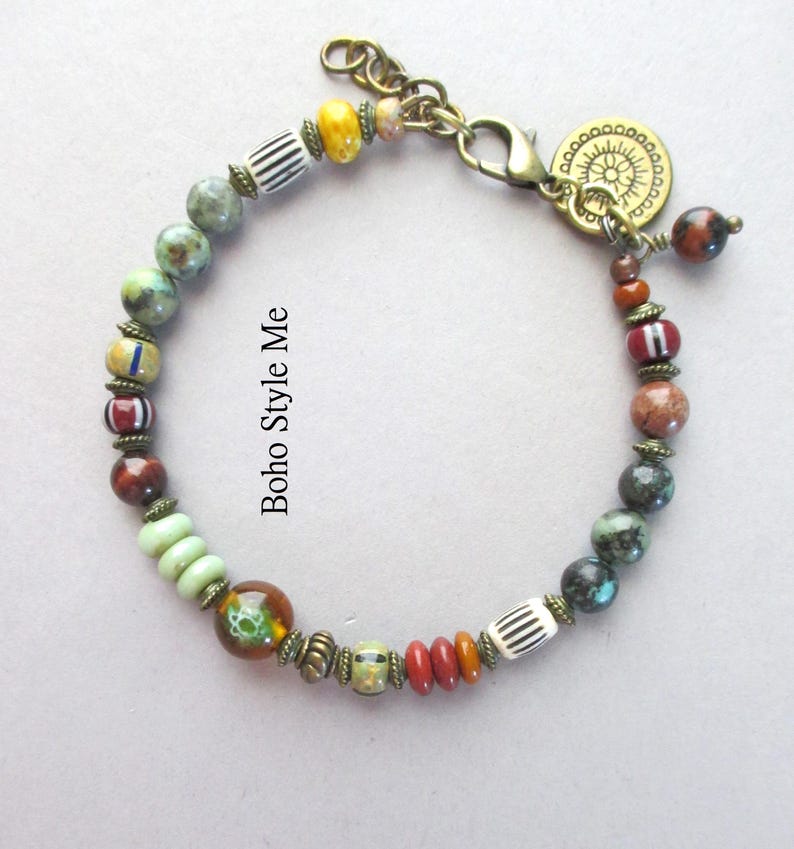 K&ouml;nnte beinhalten: Ein Perlenarmband mit einer Vielzahl bunter Perlen in Gr&uuml;n-, Braun-, Rot- und Gelbt&ouml;nen, akzentuiert durch antike Messing-Abstandshalter und einen runden Anh&auml;nger. Der Text Boho Style Me ist sichtbar. Es enth&auml;lt einen Karabinerverschluss und eine Kette.