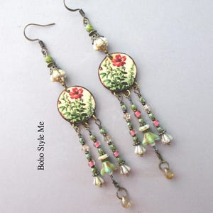 May include: A pair of dangle earrings featuring circular charms with a botanical print of red flowers and green foliage. Each earring includes bronze-toned hooks, small green and pink beads, and dangling flower-shaped charms. The text Boho Style Me is visible.