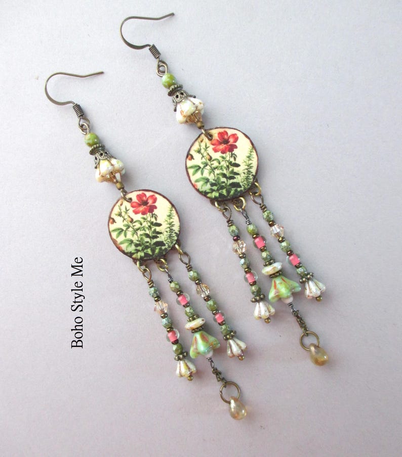 May include: A pair of dangle earrings featuring circular charms with a botanical print of red flowers and green foliage. Each earring includes bronze-toned hooks, small green and pink beads, and dangling flower-shaped charms. The text Boho Style Me is visible.