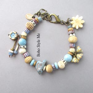 May include: A charm bracelet featuring a variety of beads in shades of blue, cream, and tan, including a bird-shaped bead, a flower-shaped charm, and a bronze-toned lobster clasp. The text Boho Style Me is printed in the center of the bracelet loop.