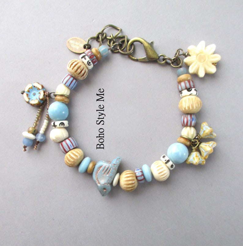 May include: A charm bracelet featuring a variety of beads in shades of blue, cream, and tan, including a bird-shaped bead, a flower-shaped charm, and a bronze-toned lobster clasp. The text Boho Style Me is printed in the center of the bracelet loop.