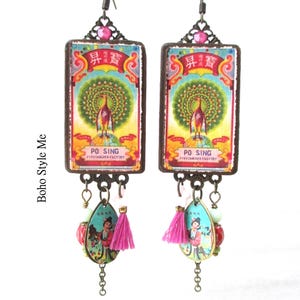 May include: A pair of rectangular drop earrings featuring a vintage-style graphic of a peacock and the text PO SING FIRECRACKER FACTORY. Below the main rectangle, each earring has a teardrop charm with a figure, a small pink tassel, and beads.