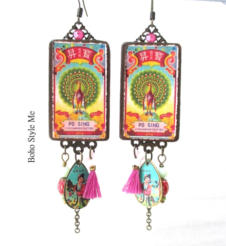 May include: A pair of rectangular drop earrings featuring a vintage-style graphic of a peacock and the text PO SING FIRECRACKER FACTORY. Below the main rectangle, each earring has a teardrop charm with a figure, a small pink tassel, and beads.