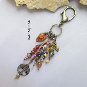 Puede incluir: Un llavero con un mosquetón y una anilla de tono bronce, adornado con múltiples hilos de cuentas de colores, pequeños dijes de hojas de metal, un dije de bellota y un colgante de árbol de la vida. El texto Boho Style Me es visible.