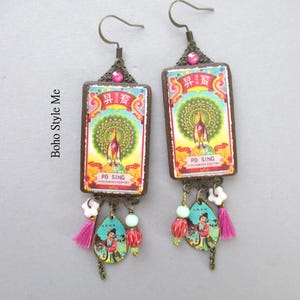 May include: A pair of rectangular drop earrings featuring a vintage Po Sing label with a peacock illustration, accented with a pink rhinestone, a small white flower bead, a light green bead, a red patterned bead, and a pink tassel.