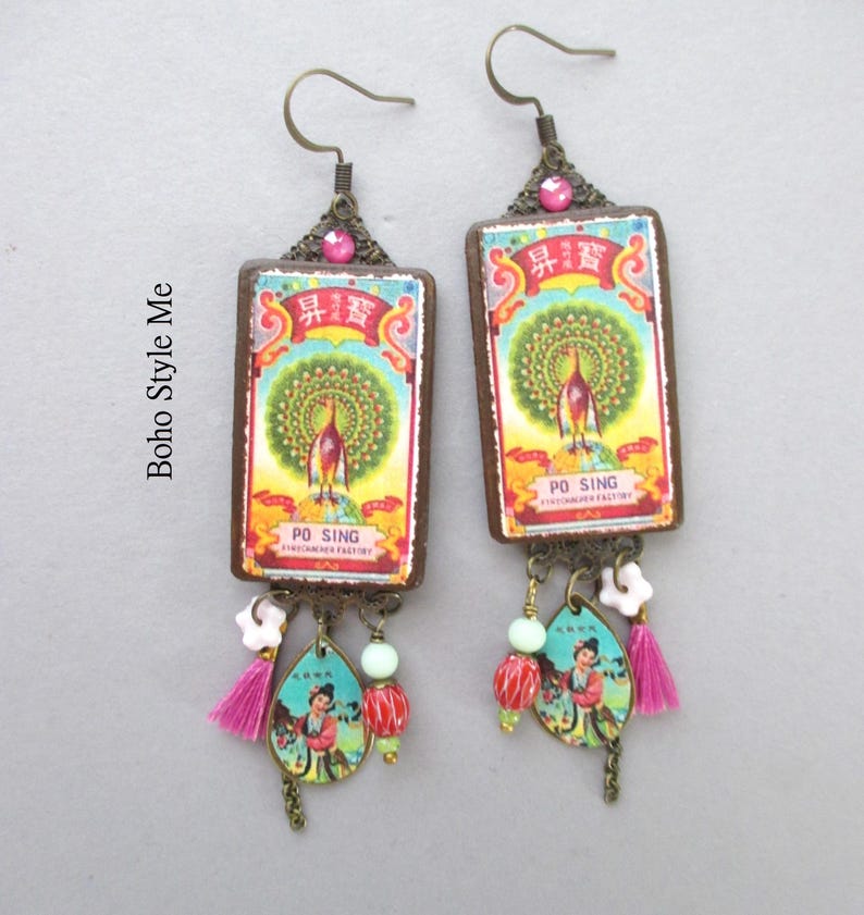 May include: A pair of rectangular drop earrings featuring a vintage Po Sing label with a peacock illustration, accented with a pink rhinestone, a small white flower bead, a light green bead, a red patterned bead, and a pink tassel.