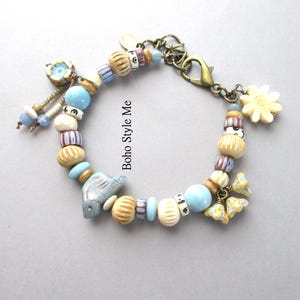 May include: A charm bracelet featuring a variety of beads in shades of blue, cream, and brown, including striped, ribbed, and solid spherical shapes. The bracelet includes a blue ceramic bird charm, floral charms, and a bronze lobster clasp.