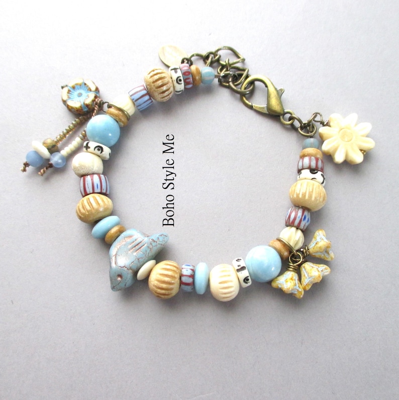 May include: A charm bracelet featuring a variety of beads in shades of blue, cream, and brown, including striped, ribbed, and solid spherical shapes. The bracelet includes a blue ceramic bird charm, floral charms, and a bronze lobster clasp.