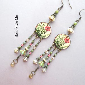 May include: A pair of dangle earrings featuring circular charms with a botanical print of red flowers and green foliage. Each earring has three beaded strands with small green, pink, and white glass beads, finished with a metallic drop. Text reads Boho Style Me.