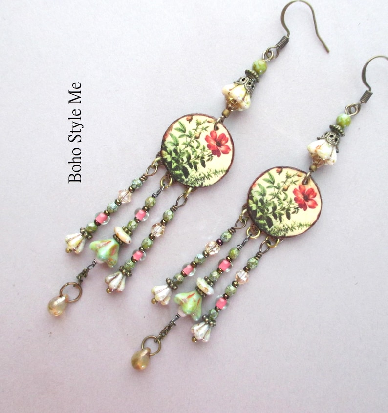 May include: A pair of dangle earrings featuring circular charms with a botanical print of red flowers and green foliage. Each earring has three beaded strands with small green, pink, and white glass beads, finished with a metallic drop. Text reads Boho Style Me.