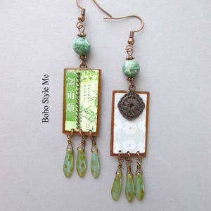 Puede incluir: Un par de pendientes colgantes con ganchos de tono cobre, cuentas redondas de color verde moteado y colgantes rectangulares. Un colgante muestra im&aacute;genes de bamb&uacute; con caracteres asi&aacute;ticos, mientras que el otro tiene un patr&oacute;n floral blanco y un medall&oacute;n.