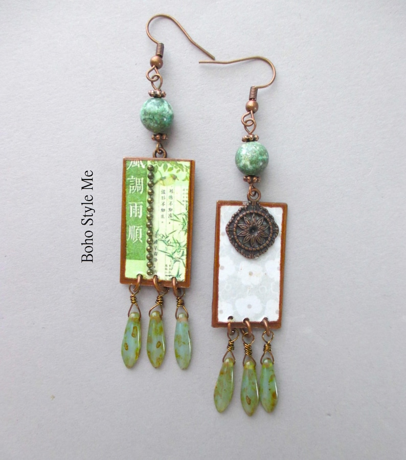 Puede incluir: Un par de pendientes colgantes con ganchos de tono cobre, cuentas redondas de color verde moteado y colgantes rectangulares. Un colgante muestra im&aacute;genes de bamb&uacute; con caracteres asi&aacute;ticos, mientras que el otro tiene un patr&oacute;n floral blanco y un medall&oacute;n.