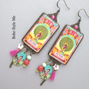 May include: A pair of rectangular drop earrings featuring a vintage-style graphic of a peacock and the text Po Sing Firecracker Factory. The earrings include bronze-toned metal hooks, pink tassels, small white flower beads, and teardrop charms.