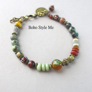 K&ouml;nnte beinhalten: Ein Perlenarmband mit einer Vielzahl von bunten Stein- und Glasperlen in Gr&uuml;n-, Rot-, Braun- und Gelbt&ouml;nen, akzentuiert durch antike Messing-Abstandshalter und einen Karabinerverschluss. Der Text Boho Style Me ist auf dem Bild zu sehen.