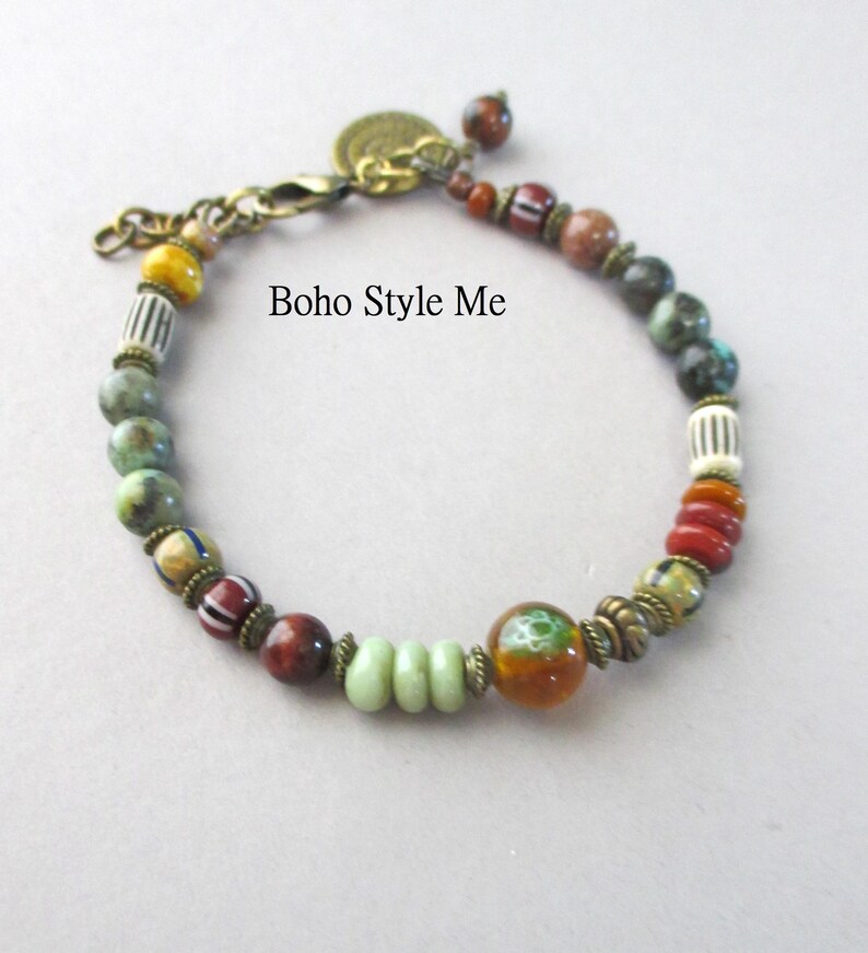 K&ouml;nnte beinhalten: Ein Perlenarmband mit einer Vielzahl von bunten Stein- und Glasperlen in Gr&uuml;n-, Rot-, Braun- und Gelbt&ouml;nen, akzentuiert durch antike Messing-Abstandshalter und einen Karabinerverschluss. Der Text Boho Style Me ist auf dem Bild zu sehen.