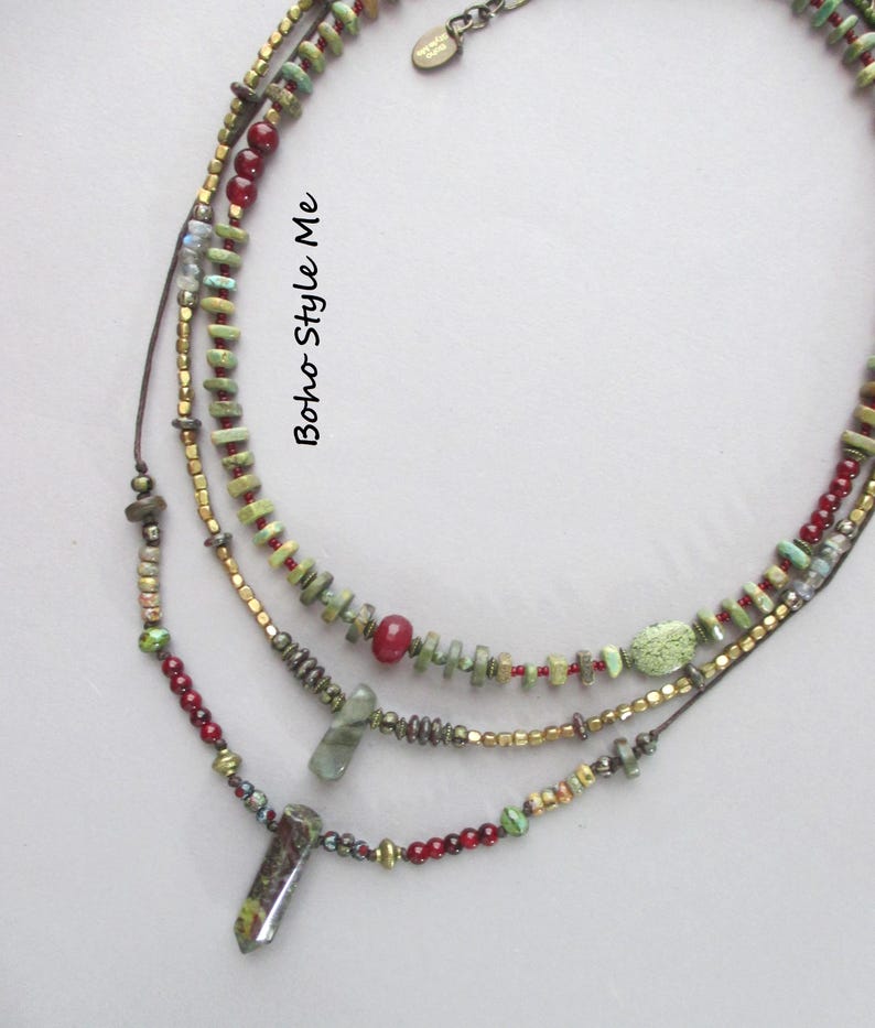 Puede incluir: Un collar de cuentas de varias hebras con cuentas verdes, rojas y doradas met&aacute;licas y dos colgantes de piedra. El collar incluye peque&ntilde;as cuentas cil&iacute;ndricas y redondas, con el texto Boho Style Me impreso verticalmente en el centro.