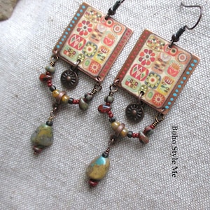Puede incluir: Un par de pendientes rectangulares con un patrón floral multicolor sobre una base de tono cobre. Cada pendiente incluye un pequeño dije en forma de sol, un alambre curvo con cuentas de colores y una cuenta similar a una piedra.