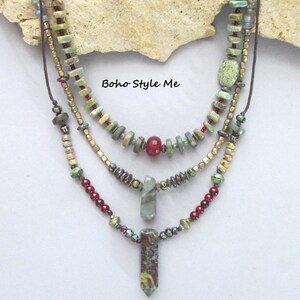 Puede incluir: Un collar de varias hebras con tres capas de cuentas en tonos verde, rojo y oro met&aacute;lico. El dise&ntilde;o incluye peque&ntilde;as piedras, cuentas redondas y dos colgantes de piedra verticales. El texto Boho Style Me es visible en el centro.