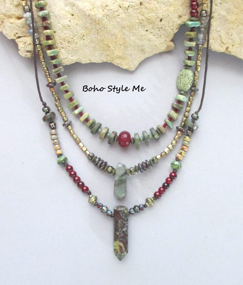 Puede incluir: Un collar de varias hebras con tres capas de cuentas en tonos verde, rojo y oro met&aacute;lico. El dise&ntilde;o incluye peque&ntilde;as piedras, cuentas redondas y dos colgantes de piedra verticales. El texto Boho Style Me es visible en el centro.