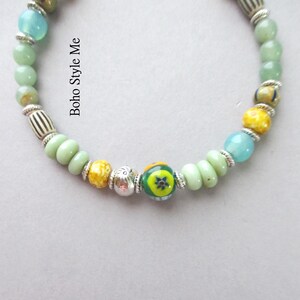 May include: A beaded bracelet featuring a variety of round and disc-shaped beads in shades of light green, yellow, and blue, interspersed with silver-toned metal spacers and a central bead with a green, yellow, and blue floral pattern. Text reads Boho Style Me.