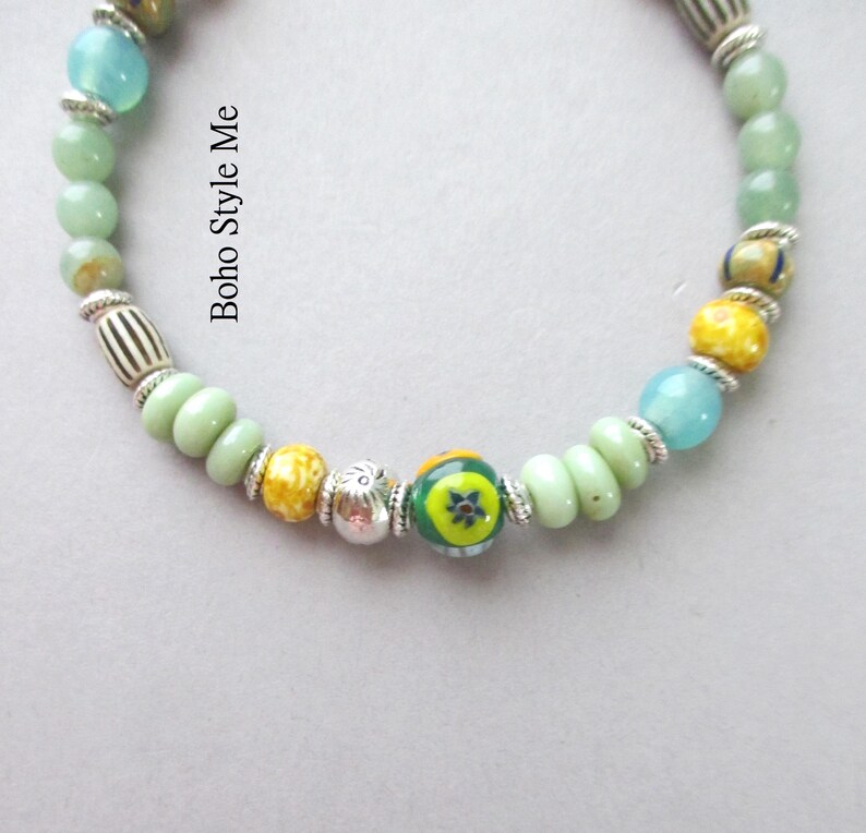 May include: A beaded bracelet featuring a variety of round and disc-shaped beads in shades of light green, yellow, and blue, interspersed with silver-toned metal spacers and a central bead with a green, yellow, and blue floral pattern. Text reads Boho Style Me.